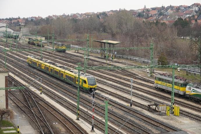 Egy többvágányos vasúti pályaudvar felülnézetből, ahol zöld-sárga személyvonat és több mozdony áll a síneken; a háttérben fák és családi házak láthatók.