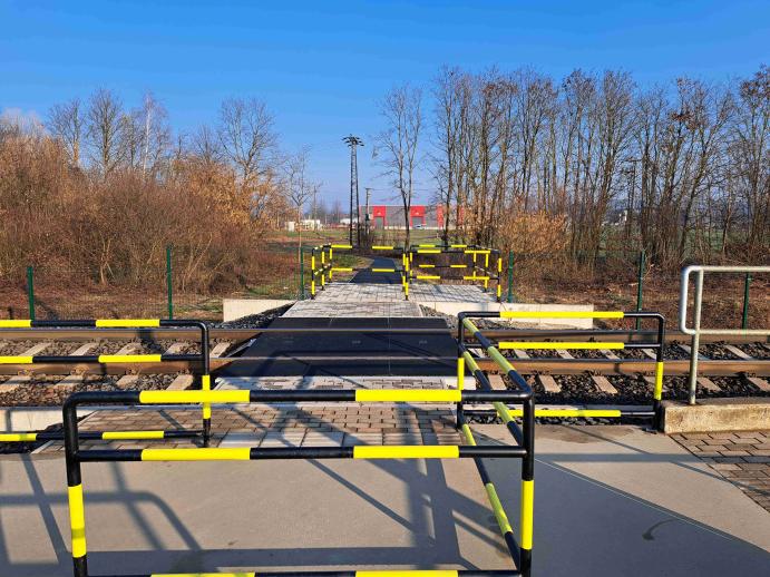 A képen egy gyalogos vasúti átjáró látható, amelyet fekete-sárga színű, cikk-cakk elrendezésű korlátok vezetnek át a síneken. A burkolt gyalogút a korlátok között halad, amelyek lassításra és körültekintő áthaladásra késztetik a gyalogosokat a vasúti pálya keresztezésekor.