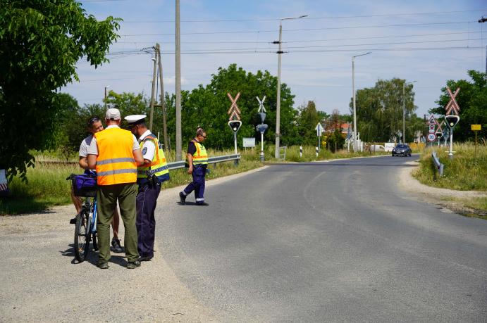 „Rendőrök és közlekedésbiztonsági szakemberek egy kerékpárossal beszélgetnek egy vasúti átjáró közelében, a biztonságos közlekedésre felhívó ellenőrzés során.