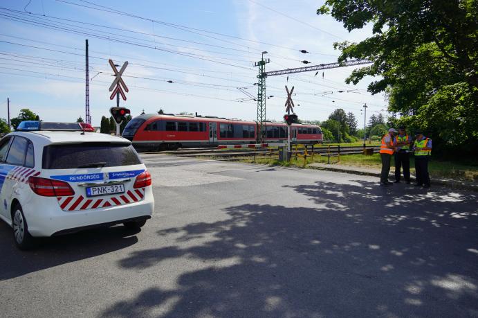 Vasúti átjáróban közlekedésbiztonsági ellenőrzés zajlik, miközben a háttérben egy személyvonat halad át.